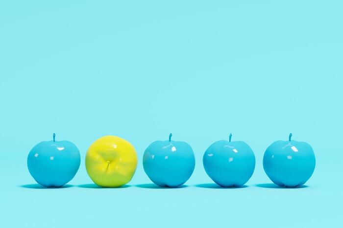Row of 4 blue apples and 1 yellow lemon in front of a light blue background, highlighting contrast of colors