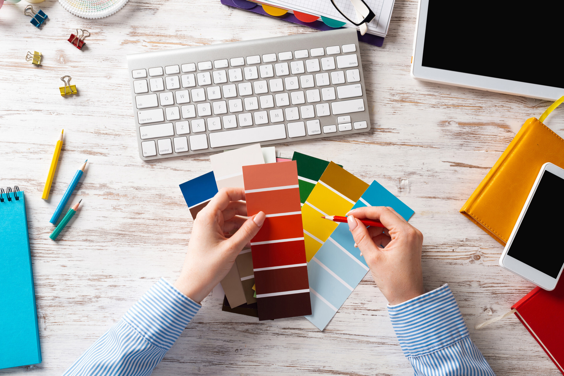 Hands comparing different paint swatches while in front of computer keyboard, mobile device, tablet, notepad, writing utensils